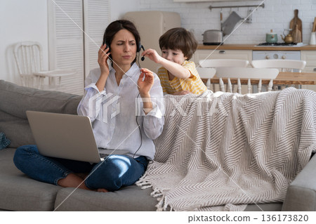 Exhausted mother sitting on couch at home, working on laptop, child distracted and asking attention Exhausted mother sitting on couch at home, working on laptop, child distracted and asking attention 136173820