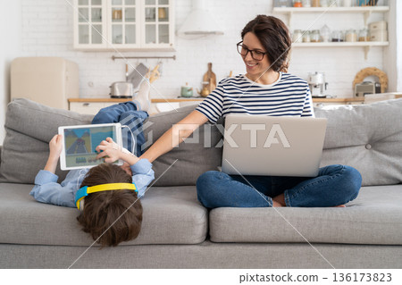 Mom freelancer remote work from home office on laptop sit on couch child playing on tablet. Lockdown 136173823