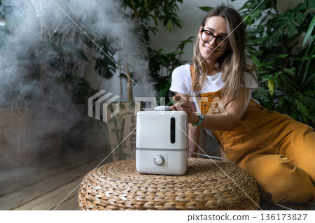 Woman gardener in overalls feeling fresh, sitting on the wooden floor, using air humidifier in home garden during heating period 136173827