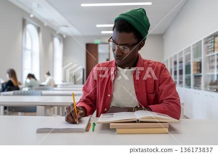 Talented young African American guy scientist wearing glasses studying in library. Education concept Talented young African American guy scientist wearing glasses studying in library. Education concept 136173837