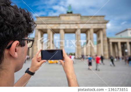Berlin, germany, august 12, 2023. Young man photographing berlin's iconic brandenburg gate with smartphone, back view capturing urban sightseeing and travel memories Berlin, germany, august 12, 2023. Young man photographing berlin's iconic brandenburg gate with smartphone, back view capturing urban sightseeing and travel memories 136173851