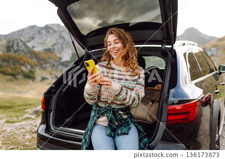 Young traveler woman sits on the rear of her car, smiling and using phone in scenic mountainous area 136173873