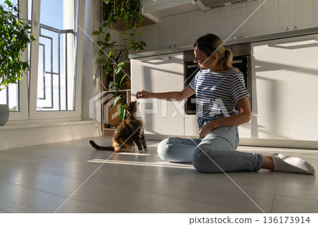 Good-natured Caucasian woman plays with kitten and has good time sits on kitchen floor near stove Good-natured Caucasian woman plays with kitten and has good time sits on kitchen floor near stove 136173914