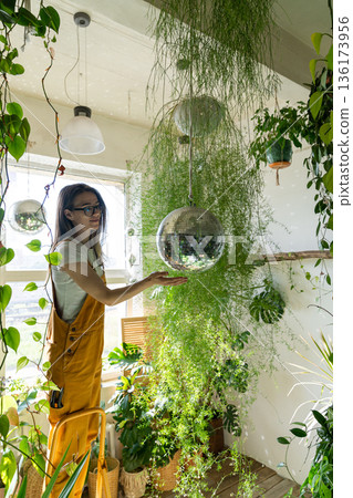 Joyful young woman gardener standing on stepladder, touches the hanging disco ball.Love of plants. Indoor garden. 136173956