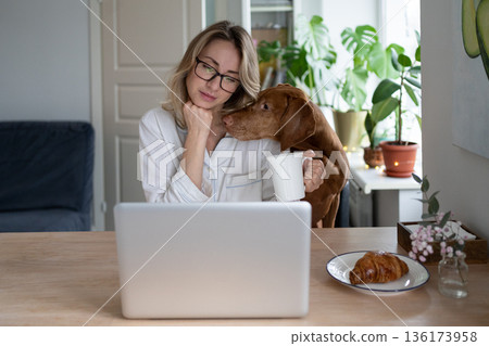 Woman in pajamas sitting at home drinking tea dog interferes with work at laptop, asks for attention 136173958