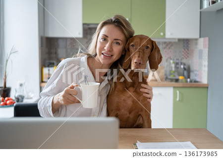 Woman in sleepwear hugging Vizsla dog, sitting on chair in kitchen, drinking tea, looking at camera 136173985