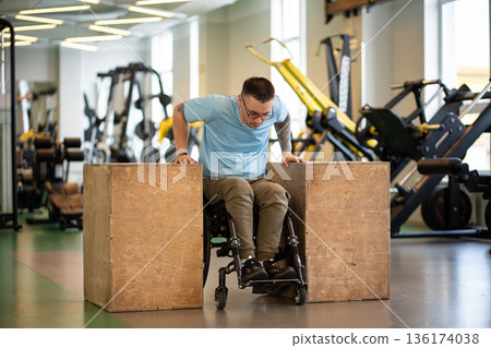 Confident man athlete in wheelchair doing push-ups strengthening exercise on wooden cubes in gym. 136174038