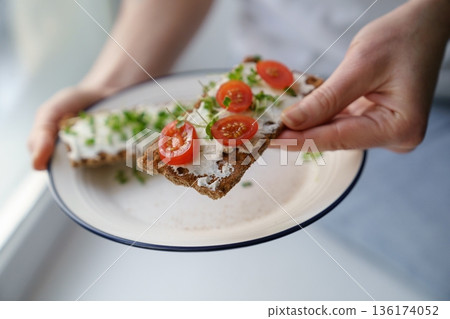 Hands holding plate with rye crisp bread with creamy cheese tofu, cherry tomato and micro greens Hands holding plate with rye crisp bread with creamy cheese tofu, cherry tomato and micro greens 136174052