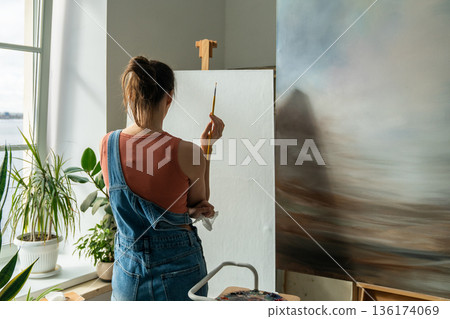 Focused female artist painting picture at home studio, holding paintbrush and looking at canvas. Focused female artist painting picture at home studio, holding paintbrush and looking at canvas. 136174069