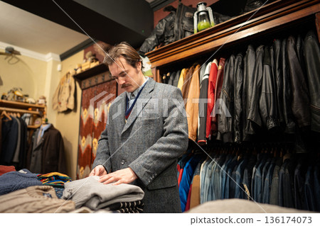 Elegant man shopping for clothing in vintage shop, choosing knitted sweaters from table display. 136174073