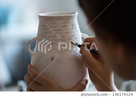 Female artisan paints text on white ceramic vase with marker on blurred background in workshop Female artisan paints text on white ceramic vase with marker on blurred background in workshop 136174124