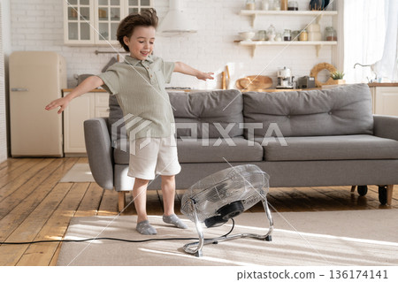 Happy smiling kid, preschool boy has fun with blowing ventilator and fresh air flow from fan at home 136174141