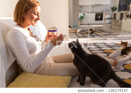 Fluffy black cat begs for food from owner eating yogurt, watches every action, sitting on floor Fluffy black cat begs for food from owner eating yogurt, watches every action, sitting on floor 136174147