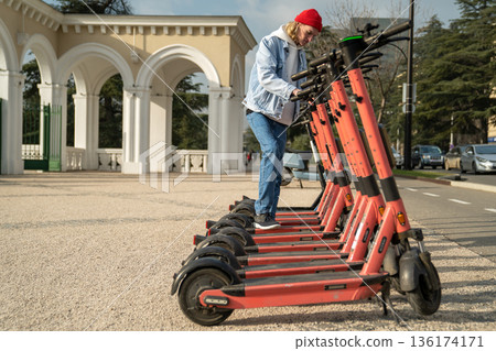 Millennial guy standing on city street unlocking e-scooter to ride around city on warm sunny day 136174171