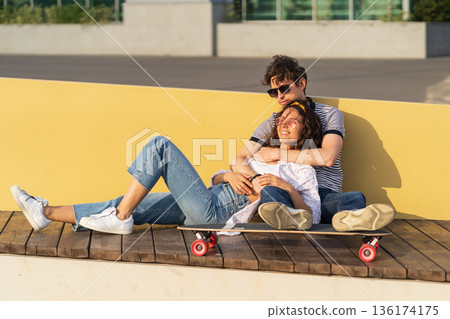 Young couple in love enjoy sunset sit hugging on longboard. Trendy man and woman chilling outdoor Young couple in love enjoy sunset sit hugging on longboard. Trendy man and woman chilling outdoor 136174175