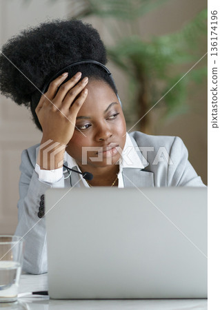 Thoughtful tired Afro businesswoman wear headphones looking at window, working at laptop, close up. 136174196