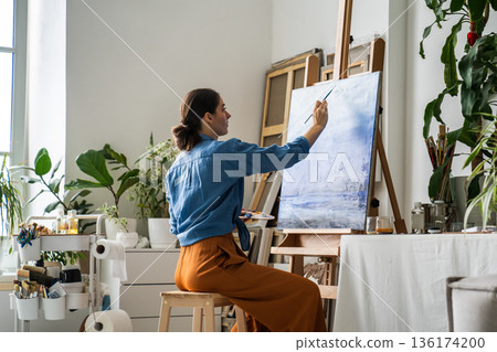 Relaxed woman artist painting picture on easel at home, sitting on chair in cozy creative atmosphere 136174200
