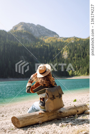 Back view young woman with backpack looking the view of the mountain lake in sunny weather. Back view young woman with backpack looking the view of the mountain lake in sunny weather. 136174227