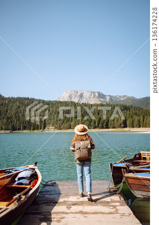 A woman with curly hair poses by the lake on a sunny day, wearing a colorful sweater and hat 136174228