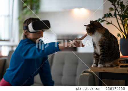 Young woman in blue sweater tries to touch domestic cat with finger wearing VR glasses in room Young woman in blue sweater tries to touch domestic cat with finger wearing VR glasses in room 136174230