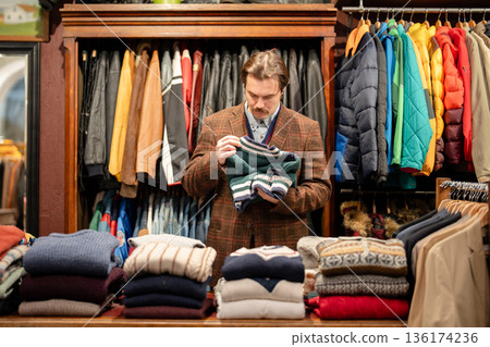 Fashionable man customer picks vintage knitwear in old fashioned second hand men's clothing boutique 136174236