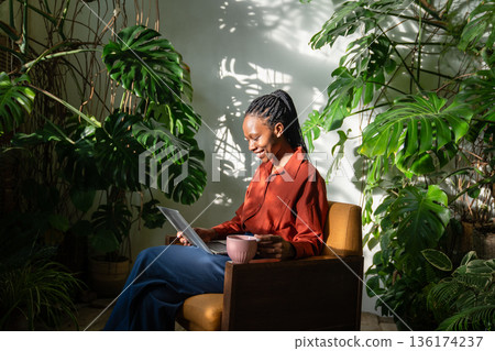 Positive African American woman relaxing in chair at home orangery filled with lush tropical plants. Positive African American woman relaxing in chair at home orangery filled with lush tropical plants. 136174237