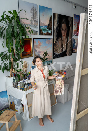Positive female artist implementing art ideas on canvas, looking at picture on easel at loft studio. Positive female artist implementing art ideas on canvas, looking at picture on easel at loft studio. 136174241