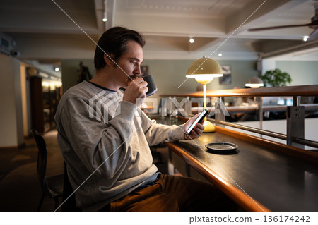 Thoughtful man enjoying coffee and scrolling social media feeds on smartphone in library cafe space. Thoughtful man enjoying coffee and scrolling social media feeds on smartphone in library cafe space. 136174242