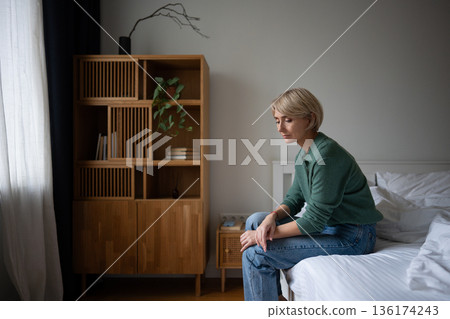 Pondering lonely woman sits on bed with upset expression, coping with thoughts about life crisis. Pondering lonely woman sits on bed with upset expression, coping with thoughts about life crisis. 136174243