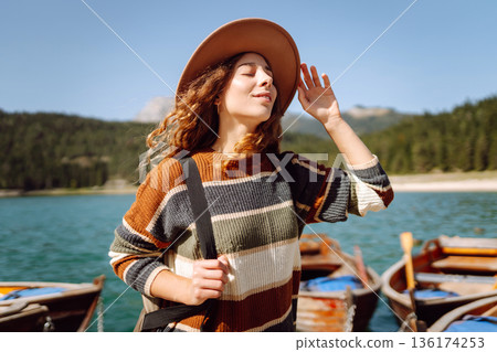 A woman with curly hair poses by the lake on a sunny day, wearing a colorful sweater and hat A woman with curly hair poses by the lake on a sunny day, wearing a colorful sweater and hat 136174253