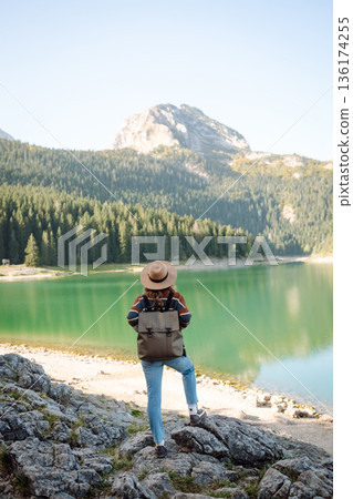 Back view young woman with backpack looking the view of the mountain lake in sunny weather. 136174255