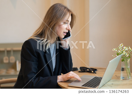 Smiling businesswoman working on laptop and talking on smartphone during lunch break in cafe. Smiling businesswoman working on laptop and talking on smartphone during lunch break in cafe. 136174263