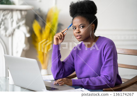 Afro-American millennial student woman with afro hairstyle looking at camera while making researches browsing information on laptop, preparing for exam online, holding a pen. Afro-American millennial student woman with afro hairstyle looking at camera while making researches browsing information on laptop, preparing for exam online, holding a pen. 136174267