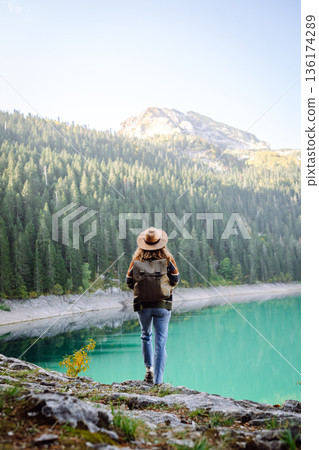 Back view young woman with backpack looking the view of the mountain lake in sunny weather. Back view young woman with backpack looking the view of the mountain lake in sunny weather. 136174289