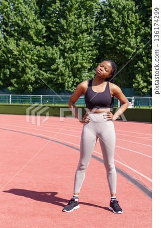 African American sportswoman standing on red running track does stretching in sunny morning African American sportswoman standing on red running track does stretching in sunny morning 136174299