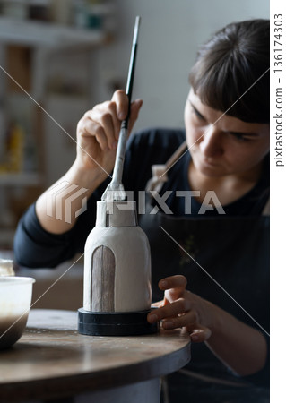 Happy concentrated young craftswoman paints clay vase sitting at work table closeup Happy concentrated young craftswoman paints clay vase sitting at work table closeup 136174303