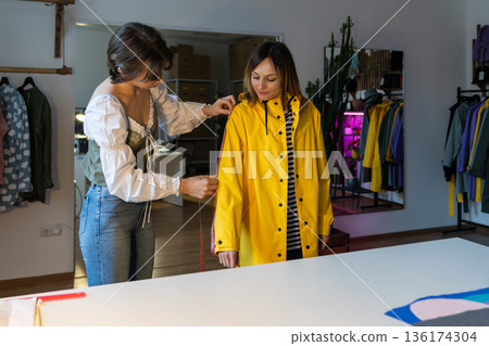 Fashion designer workshop with female tailor measuring and fitting bespoke yellow rain jacket. Fashion designer workshop with female tailor measuring and fitting bespoke yellow rain jacket. 136174304