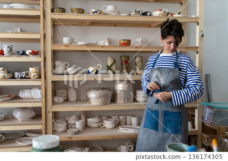 Woman ceramic artist tying apron at workplace in pottery studio. Female potter in ceramics workshop Woman ceramic artist tying apron at workplace in pottery studio. Female potter in ceramics workshop 136174305