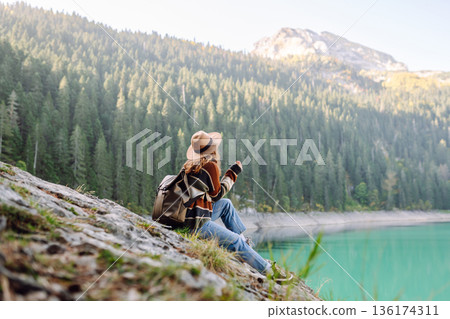 Back view young woman with backpack looking the view of the mountain lake in sunny weather. 136174311