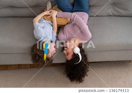 Happy mom and kid lying upside down on couch at home, laughing, tickle each other listening to music 136174313