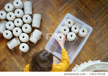 Top view of woman freelancer holding white macrame cotton ropes in skeins, puts working material in a plastic box. Crochet, weaving crafts Top view of woman freelancer holding white macrame cotton ropes in skeins, puts working material in a plastic box. Crochet, weaving crafts 136174320