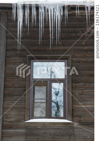 Ice stalactite hanging from the roof with wooden wall. Poor thermal insulation of the roof. Winter season Ice stalactite hanging from the roof with wooden wall. Poor thermal insulation of the roof. Winter season 136174321