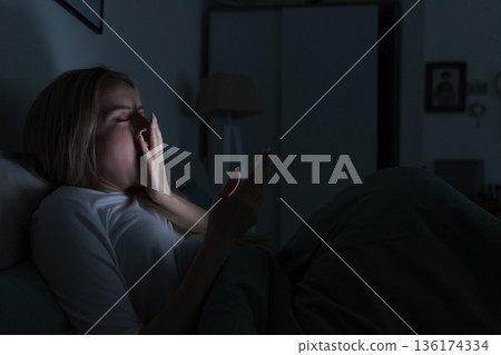 Sleepy tired woman lying in bed, using smartphone, covering mouth with hand, yawning. Insomnia Sleepy tired woman lying in bed, using smartphone, covering mouth with hand, yawning. Insomnia 136174334