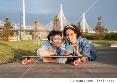 Young stylish girl show guy text message on smartphone lying on longboard in urban park outdoors Young stylish girl show guy text message on smartphone lying on longboard in urban park outdoors 136174335