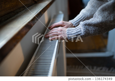 Woman in grey sweater puts hands on room central heating battery to warm up in cold apartment Woman in grey sweater puts hands on room central heating battery to warm up in cold apartment 136174349