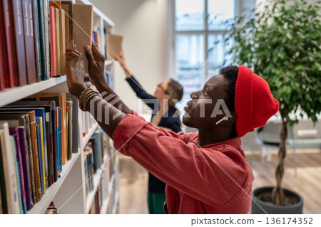 Focused puzzled on finding right educational literature student looks through bookshelves in library Focused puzzled on finding right educational literature student looks through bookshelves in library 136174352