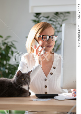 Woman working on computer from home office, talking on mobile phone, selective focus on sleepy black cat nearby on the table Woman working on computer from home office, talking on mobile phone, selective focus on sleepy black cat nearby on the table 136174463