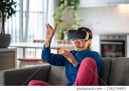 Young woman sitting on comfortable sofa plays games in VR glasses and explores virtual world Young woman sitting on comfortable sofa plays games in VR glasses and explores virtual world 136174465