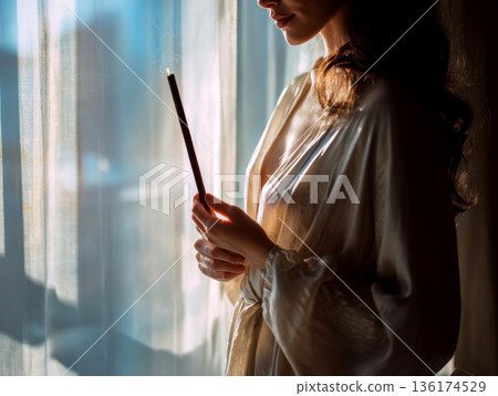 A woman stands by a window holding a lit Palo Santo stick. Soft light comes through the curtains. She is wearing a robe and appears focused on the smoke 136174529
