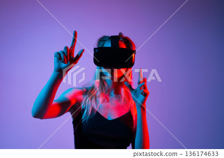 Female gamer adjusting VR headset while playing virtual reality game in neon red blue lighting studio environment 136174633